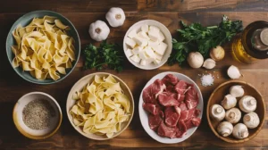 Overhead view of raw ingredients for beef stroganoff including beef strips, mushrooms, egg noodles, onions, sour cream, and herbs.